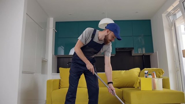 Handsome bearded man dressed in workwear cleaning the floor in the cuisine using the mop and dancing