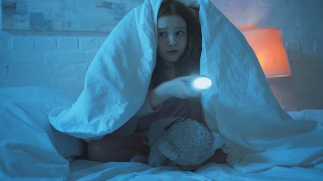 Scared Child Sitting Under Blanket With Flashlight At Night