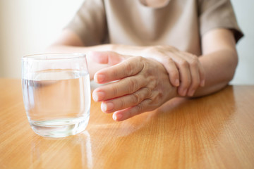 Elderly woman hands w/ tremor symptom reaching out for a glass of water on wood table. Cause of...