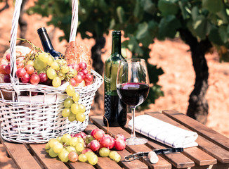 glass of red wine grapes and picnic basket on table in field