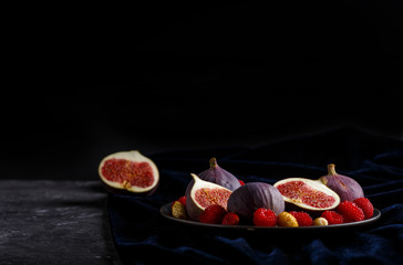 Fresh figs, strawberries and raspberries on blue plate on black concrete background and blue velvet textile. side view, selective focus, low key