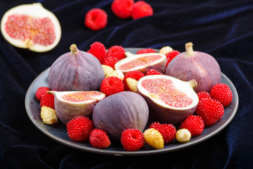 Fresh figs, strawberries and raspberries on blue plate on black concrete background and blue velvet textile. side view, selective focus