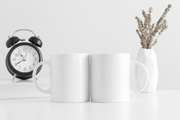 Two white mugs mockup with a lavender in a vase, book and a clock on a white table.
