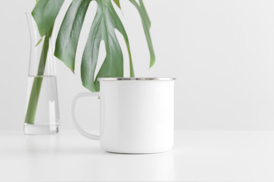 Enamel mug mockup with a monstera leaf in a glass vase on a white table.