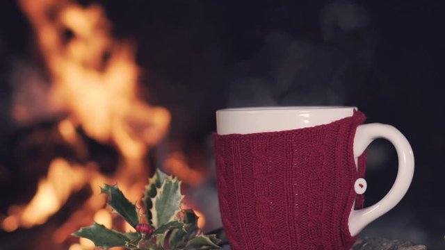 Winter, cup of hot coffee with steam on a wooden table close to a fireplace and a holly. Slow motion. 