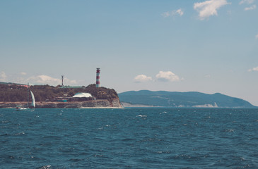 Fototapeta premium View of the coastal lighthouse on a cloudless day. The yacht drifting near a cliff on a boat trip.