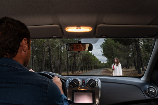 Scared man driving his car on a forest road and looking at a female ghost through the front windshield. Halloween nightmare. Halloween horror scene.