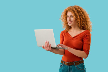smiling redhead woman using laptop isolated on blue