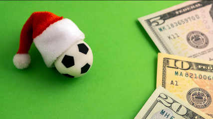 Soccer ball in santa hat next to us dollar bills on a green background. The concept of sports betting, cash incentives for football players, corruption and approval of financial in the new year.
