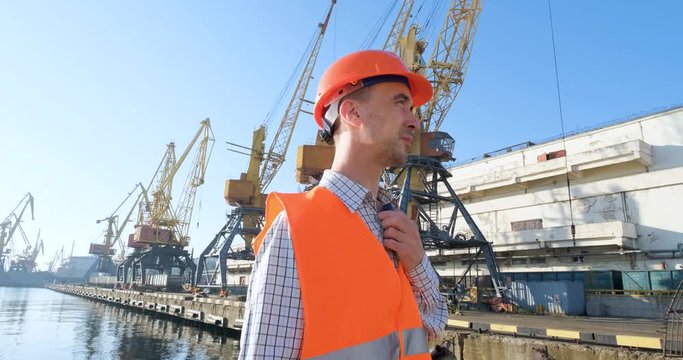 male worker of sea harbor in orange helmet and safety west, cranes and sea background 4K UHD futage 	