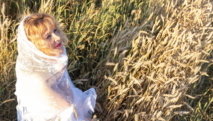 A girl with red hair is sitting on a field with ripe ears of corn. A woman in a white scarf, veil, looks at the sunset and rejoices. Countryside and sunset.