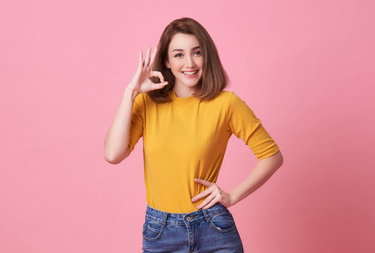 Excited beautiful woman showing OK gesture, looking at camera  wearing casual yellow t-shirt isolated on pink background.