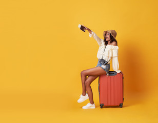 Cheerful young african woman dressed in summer clothes sitting on a suitcase and pointing finger at copy space isolated over yellow background.