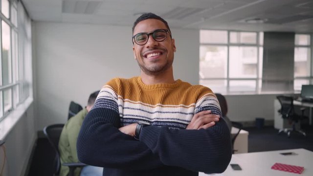 African young businessman standing in office with his arms crossed looking at camera with diverse colleagues talking in background