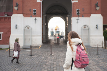 Fototapeta na wymiar tourist young girl in autumn jacket with backpack looking at Kremlin gates standing on Red Square with old woman in headscarf walking by on paving