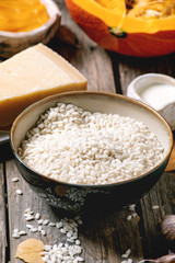 Ingredients for cooking pumpkin risotto. Raw uncooked risotto rice in ceramic bowl, sliced pumpkin, cream, seeds, parmesan cheese, garlic and autumn leaves over dark wooden background. Close up