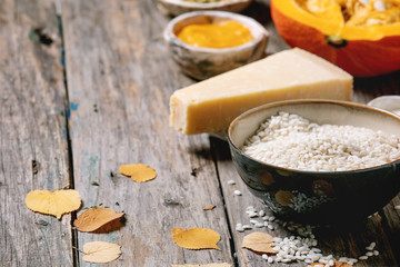 Ingredients for cooking pumpkin risotto. Raw uncooked risotto rice in ceramic bowl, sliced pumpkin, cream, seeds, parmesan cheese, garlic and autumn leaves over dark wooden background. Close up
