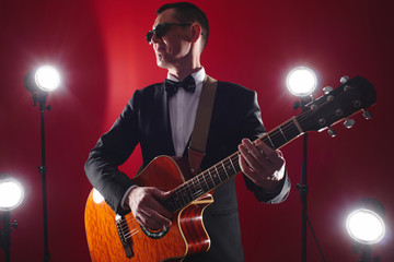 Obraz na płótnie Canvas Portrait of classical musician with guitar in red studio with stage lighting. Guitarist in black glasses and suit with a bow tie improvises on instrument