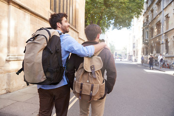 Rear View Of Male Gay Couple On Vacation Wearing Backpacks Walking Along City Street