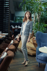 full length portrait of beautiful young smiling business brunette woman in elegant gray pantsuit and white shirt stands in a restaurant