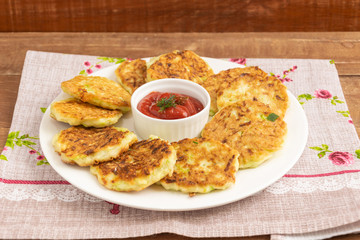 Fritters lie on a white plate. In the middle is a gravy boat with tomato sauce. A dish stands on a wooden table covered by a napkin. Fritters made from zucchini.