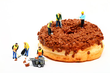 Conceptual image of a miniature figure workmen stood on a chocolate doughnut making the hole in the centre while cleaners tidy up the fallen toppings