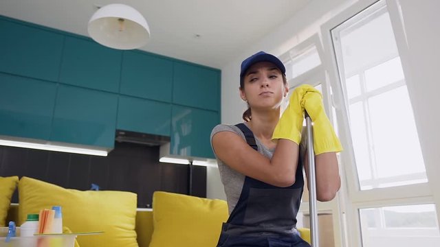 Sad young woman of the cleaning service in yellow rubber gloves holding a mop and sitting on the comfortable sofa in the middle of the kitchen