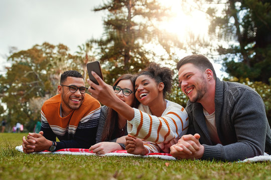 Smiling multi ethnic group of friends lying on green grass smiling while taking selfie on smartphone in the park - very green nature