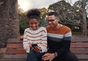 Portrait of an african young couple siting together on bench in park looking at mobile phone