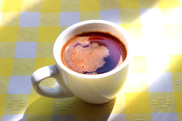 Cup of foamy morning coffee on a yellow tablecloth in the sunlight.