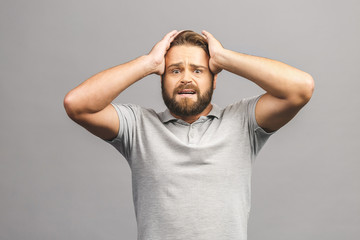 Upset unhappy young man in casual squeezing head with hands, suffering from headache. People, stress, tension and migraine concept isolated against grey background.