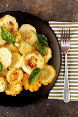 Fried zucchini in a black plate, sliced ​​vegetables. Seasonal vegetable meal, fork and striped napkin. Delicious healthy autumn food. Dark concrete background. Flat lay, copy space.