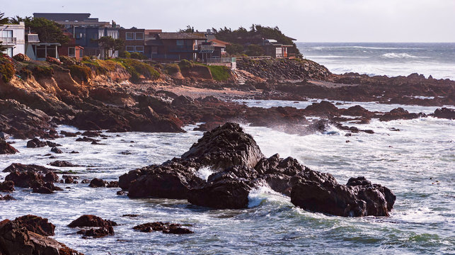 expensive beach homes sit dangerously close to the edge of a cliff above a rocky beach on the pacific coast near cambria in california