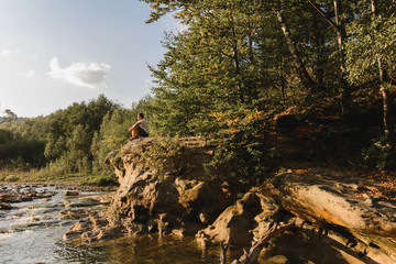 A young man sits on the edge of a stone near the river, watching the sunset.