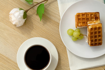 Breakfast. Cup of black coffee with Belgian waffles on wooden background top view and white flower..