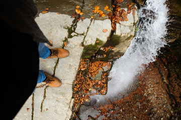 man legs in blue jeans and brown boots sitting on the edge looking at waterfall