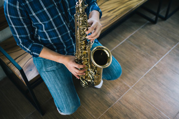 Obraz premium A young man sitting on wooden chair and plays saxophone in modern studio with window light, music background with copy space.