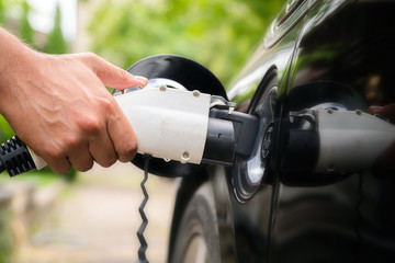 Mans hand inserting charger plug into electric car in green environment background. New energy vehicle, NEV is being loaded with electricity power. Ecology, modern day cars