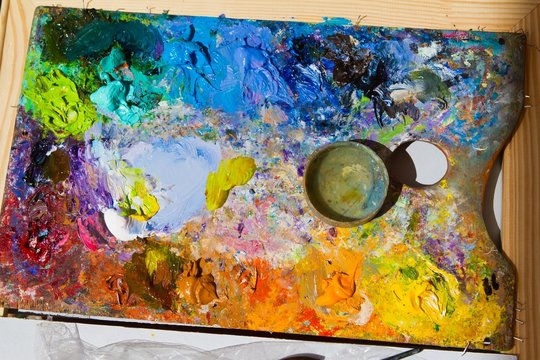 painter's wooden palette with mixed fresh bright oil paints, in disorder lays on wooden easel, summer plainair painting session, artistic flatlay in bright sunshine