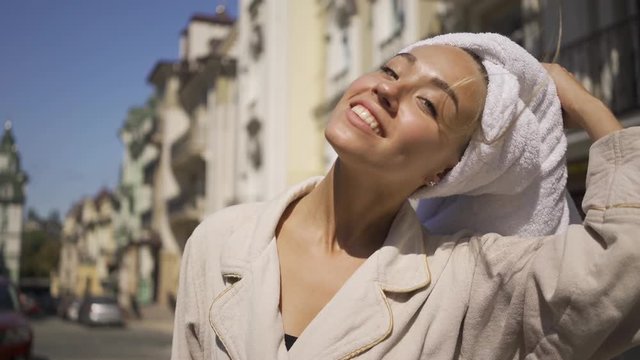 Portrait of happy confident young woman in bathrobe with towel on head walking down the street drinking coffee, turning and looking at camera smiling. The girl taking off towel from her hair