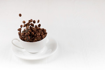 A cup with coffee beans in the air flying in flight like a spray. On background of a white wooden table, coffee shop advertising concept, copy space.