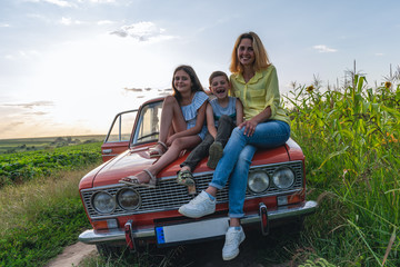 Family vacation, mom with theyr teen children all in casual clothes sitting together on a hood of the retro car among the field, all are happy and smiling