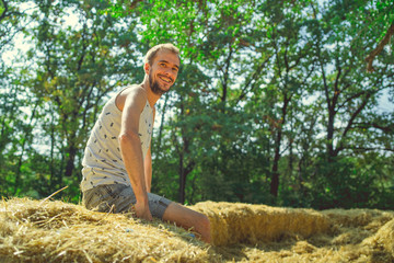 Naklejka na ściany i meble A young handsome man with beard of slim build sits in the hay and holds hay in his hands against the background of green trees in the park.