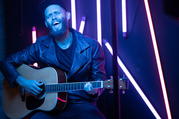 Obraz premium Singer and guitar player singing on a stage with neon lights. He is a rocker and he is wearing leather biker jacket or asymmetric zip jacket with black cap, jeans