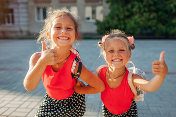 Happy sisters girls wearing backpacks and showing thumbs up. Kids pupils having fun outdoors school building.