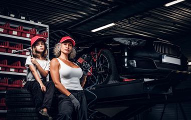 Naklejka na ściany i meble Experienced serious woman and her little helper are posing for photographer at dark auto service as great team.