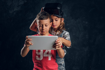 Little boy and girl are focused on tablet, girl has vr goggles on head.