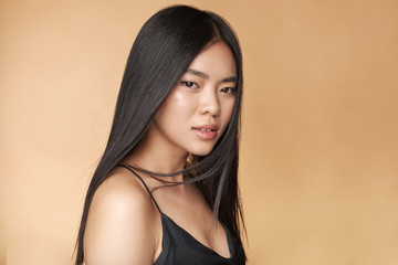 A young beautiful asian girl dressed in a black dress with natural makeup and shiny hair in the wind stands on a beige yellow natural background. Cosmetics concept. Shampoo.