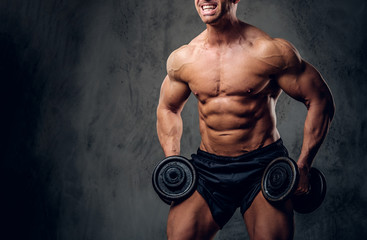 Happy smiling bodybuilder is doing exercises with barbels and showing his muscules on the dark background.