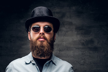 Portrait of groomed bearded man in hat and sunglasses over dark background.